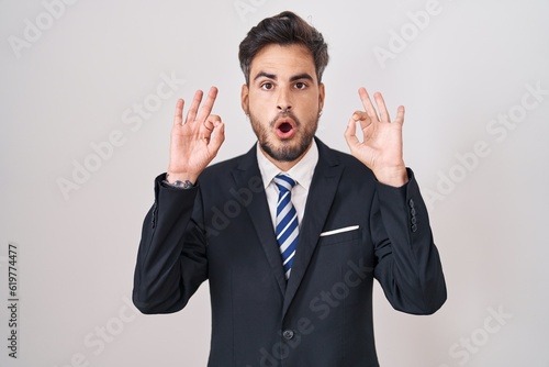 Young hispanic man with tattoos wearing business suit and tie looking surprised and shocked doing ok approval symbol with fingers. crazy expression