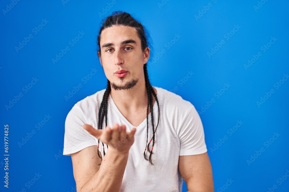 Hispanic man with long hair standing over blue background looking at the camera blowing a kiss with hand on air being lovely and sexy. love expression. Hispanic man with long hair standing over blue background looking at the camera blowing a kiss with hand on air being lovely and sexy. love expression.