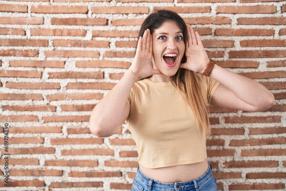 Young brunette woman standing over bricks wall smiling cheerful playing peek a boo with hands showing face. surprised and exited Young brunette woman standing over bricks wall smiling cheerful playing peek a boo with hands showing face. surprised and exited