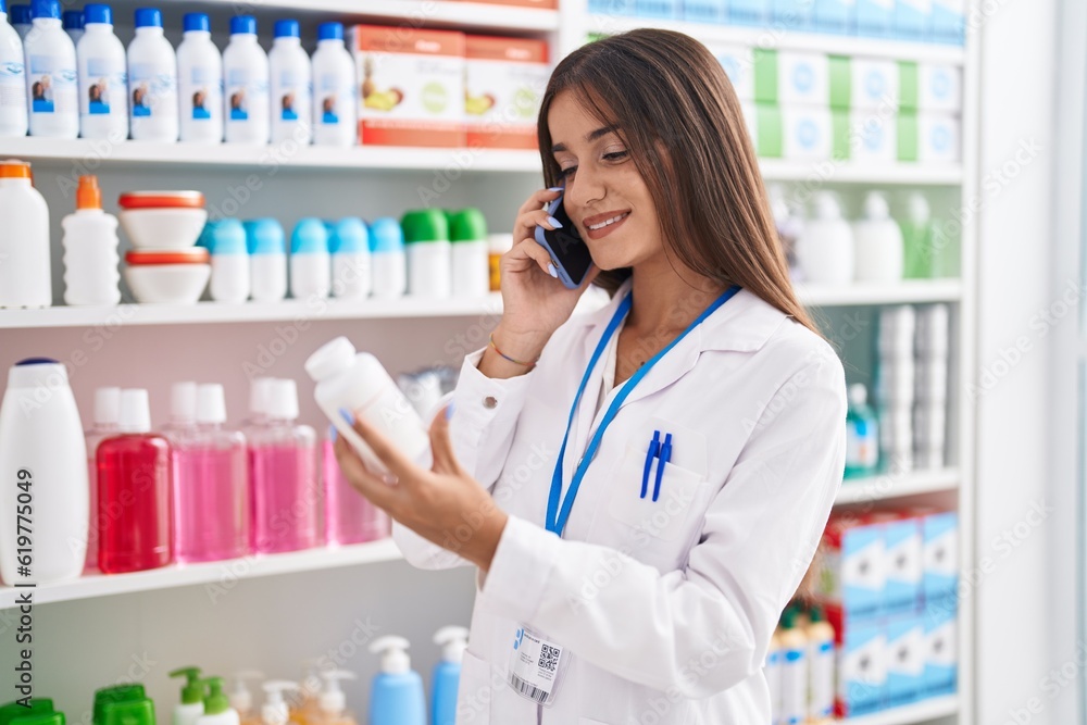 Young beautiful hispanic woman pharmacist holding pills bottle talking on smartphone at pharmacy