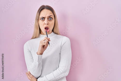 Young blonde woman standing holding pen over pink background in shock face, looking skeptical and sarcastic, surprised with open mouth