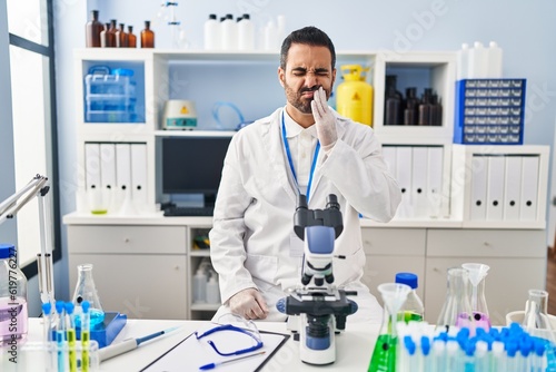 Young hispanic man with beard working at scientist laboratory touching mouth with hand with painful expression because of toothache or dental illness on teeth. dentist