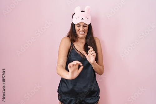 Young brunette woman wearing sleep mask and pyjama disgusted expression, displeased and fearful doing disgust face because aversion reaction. with hands raised