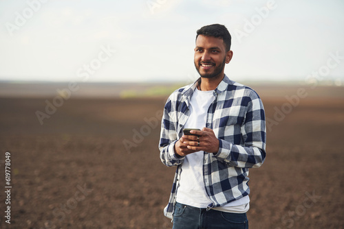 Smartphone in hands. Cultivated agricultural field. Handsome Indian man