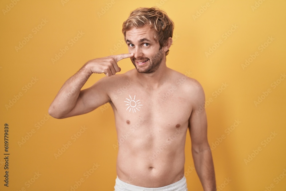 Caucasian man standing shirtless wearing sun screen pointing with hand finger to face and nose, smiling cheerful. beauty concept