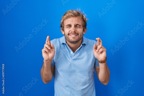 Caucasian man standing over blue background gesturing finger crossed smiling with hope and eyes closed. luck and superstitious concept. © Krakenimages.com