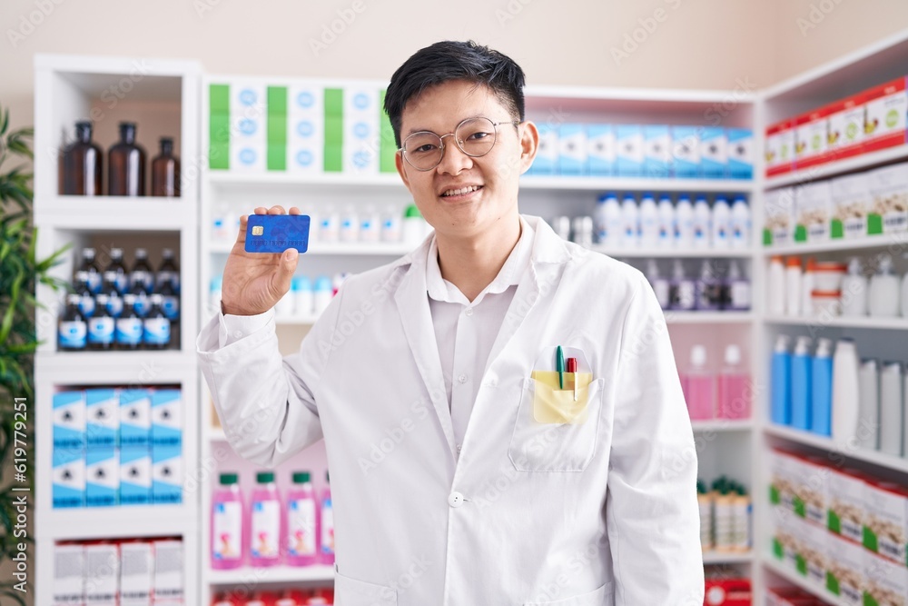Young asian man working at pharmacy drugstore holding credit card looking positive and happy standing and smiling with a confident smile showing teeth