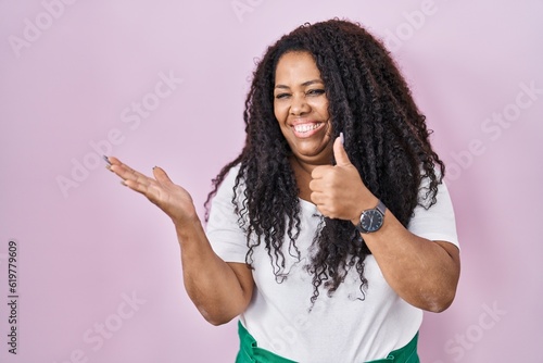 Plus size hispanic woman standing over pink background showing palm hand and doing ok gesture with thumbs up, smiling happy and cheerful