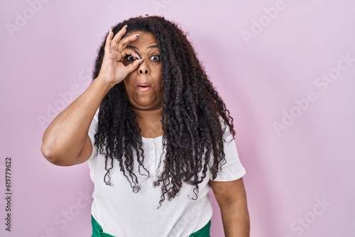Plus size hispanic woman standing over pink background doing ok gesture shocked with surprised face, eye looking through fingers. unbelieving expression.