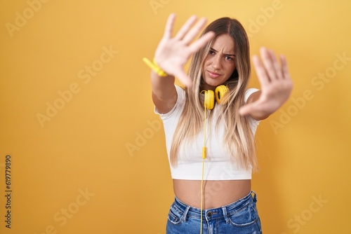 Young blonde woman standing over yellow background wearing headphones doing frame using hands palms and fingers, camera perspective