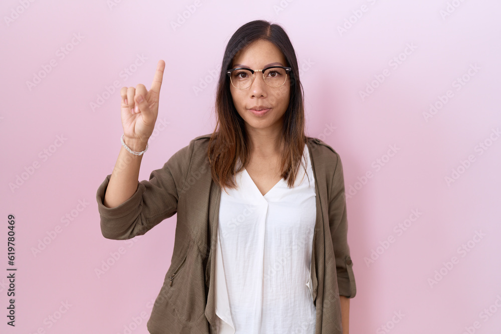 Middle age chinese woman wearing glasses over pink background showing and pointing up with finger number one while smiling confident and happy. Middle age chinese woman wearing glasses over pink background showing and pointing up with finger number one while smiling confident and happy.