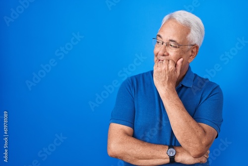 Middle age man with grey hair standing over blue background looking stressed and nervous with hands on mouth biting nails. anxiety problem.