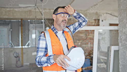 Middle age man builder holding hardhat sweating at construction site