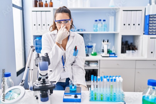Young hispanic woman working at scientist laboratory smelling something stinky and disgusting, intolerable smell, holding breath with fingers on nose. bad smell