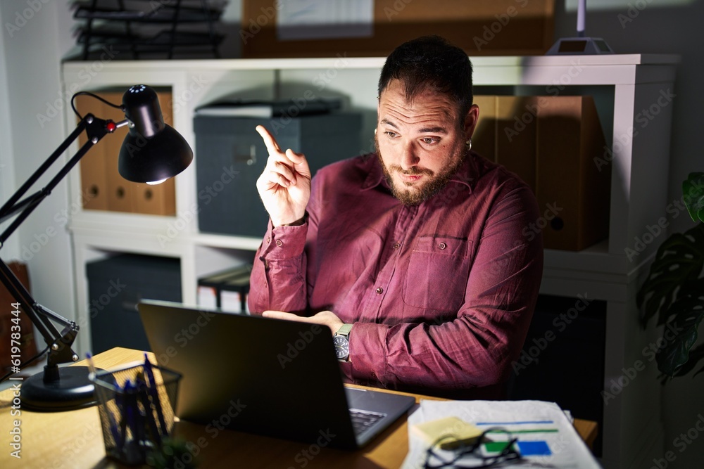 Plus size hispanic man with beard working at the office at night with a big smile on face, pointing with hand and finger to the side looking at the camera. Plus size hispanic man with beard working at the office at night with a big smile on face, pointing with hand and finger to the side looking at the camera.
