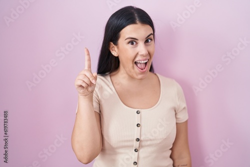 Young hispanic woman standing over pink background pointing finger up with successful idea. exited and happy. number one.