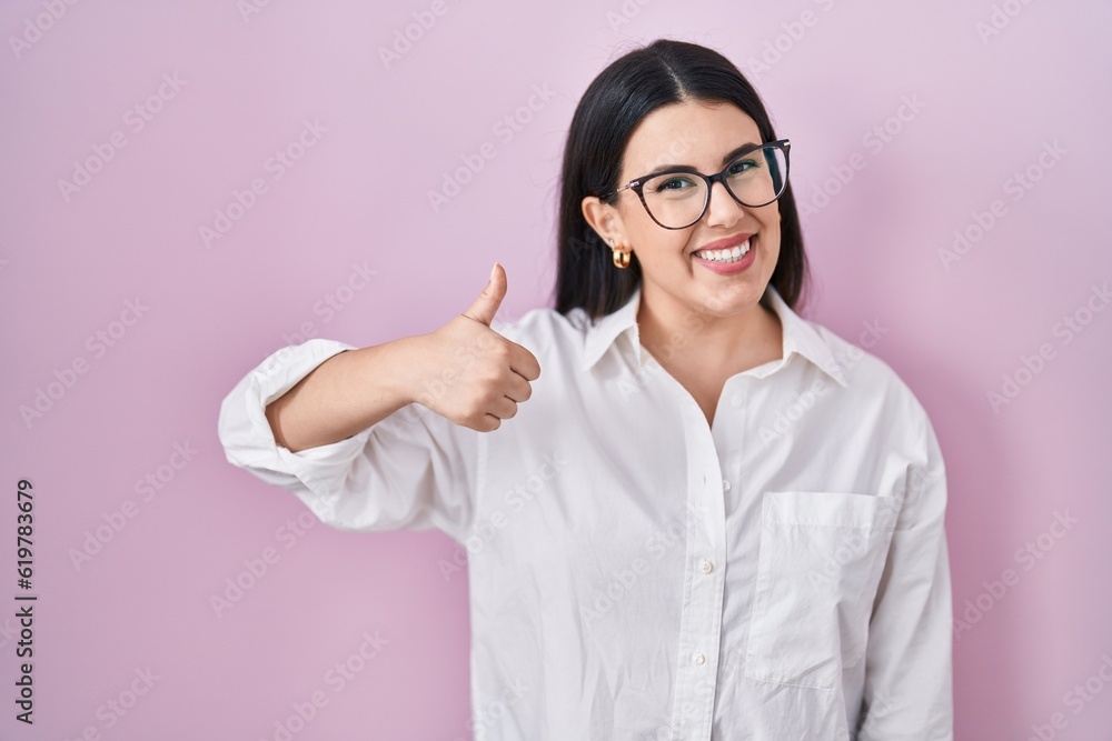 Young brunette woman standing over pink background doing happy thumbs up gesture with hand. approving expression looking at the camera showing success. Young brunette woman standing over pink background doing happy thumbs up gesture with hand. approving expression looking at the camera showing success.