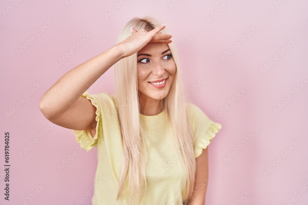 Caucasian woman standing over pink background very happy and smiling looking far away with hand over head. searching concept.