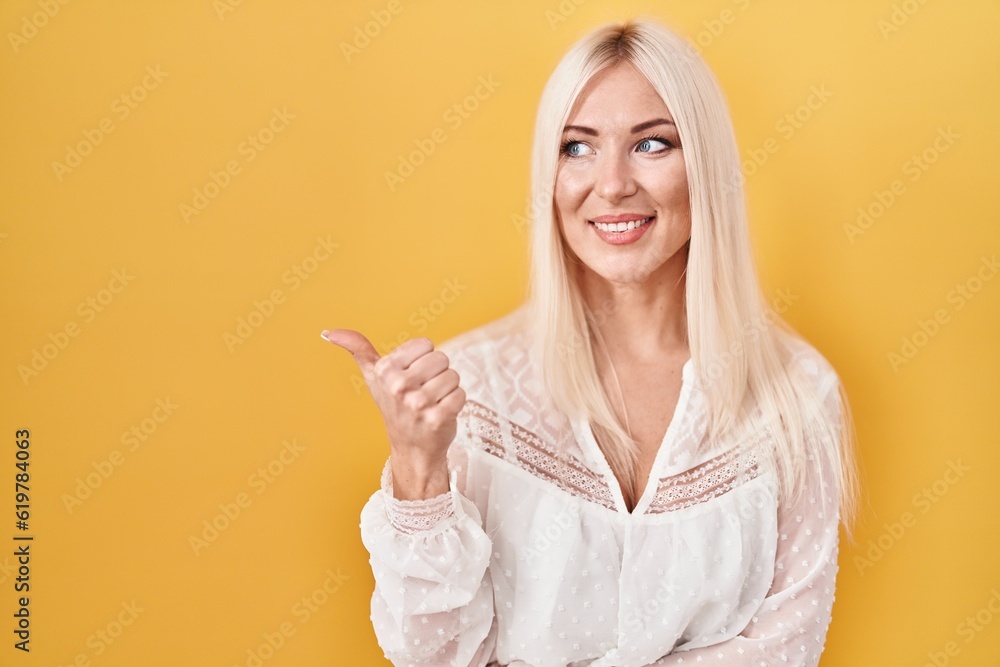 Caucasian woman standing over yellow background smiling with happy face looking and pointing to the side with thumb up. Caucasian woman standing over yellow background smiling with happy face looking and pointing to the side with thumb up.