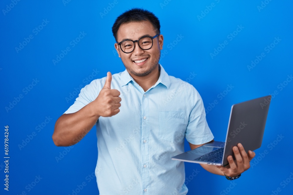 Chinese young man using computer laptop doing happy thumbs up gesture with hand. approving expression looking at the camera showing success. Chinese young man using computer laptop doing happy thumbs up gesture with hand. approving expression looking at the camera showing success.