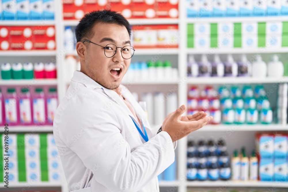 Chinese young man working at pharmacy drugstore pointing aside with hands open palms showing copy space, presenting advertisement smiling excited happy Chinese young man working at pharmacy drugstore pointing aside with hands open palms showing copy space, presenting advertisement smiling excited happy