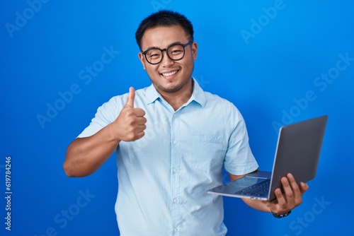Chinese young man using computer laptop doing happy thumbs up gesture with hand. approving expression looking at the camera showing success.