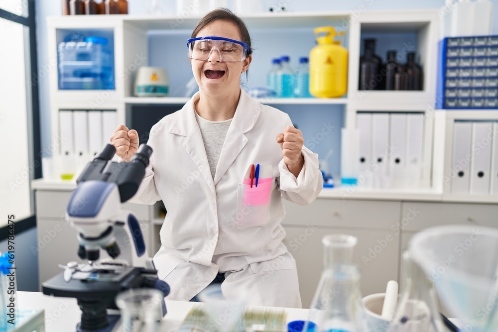 Hispanic girl with down syndrome working at scientist laboratory excited for success with arms raised and eyes closed celebrating victory smiling. winner concept. Hispanic girl with down syndrome working at scientist laboratory excited for success with arms raised and eyes closed celebrating victory smiling. winner concept.