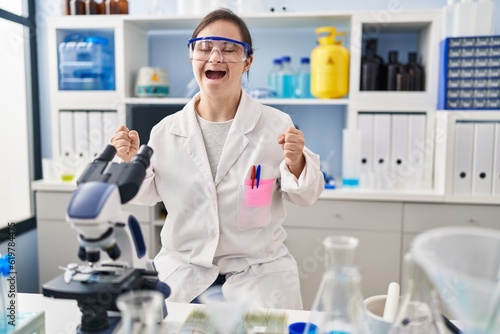 Hispanic girl with down syndrome working at scientist laboratory excited for success with arms raised and eyes closed celebrating victory smiling. winner concept.