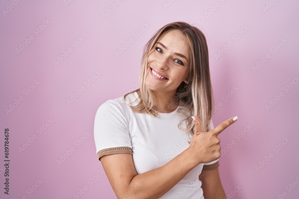 Blonde caucasian woman standing over pink background cheerful with a smile on face pointing with hand and finger up to the side with happy and natural expression Blonde caucasian woman standing over pink background cheerful with a smile on face pointing with hand and finger up to the side with happy and natural expression