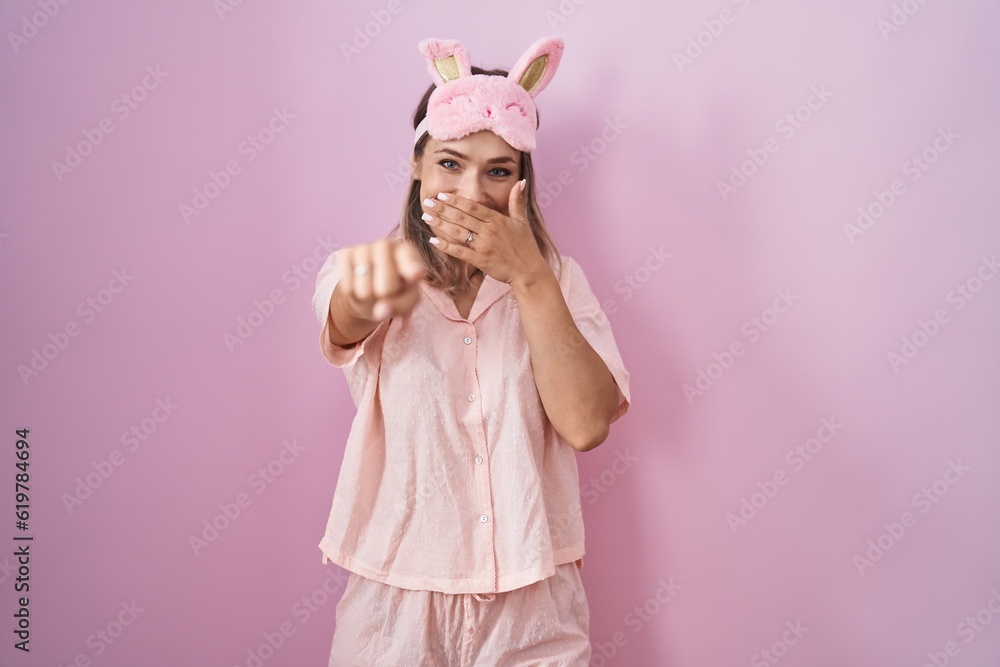 Blonde caucasian woman wearing sleep mask and pajama laughing at you, pointing finger to the camera with hand over mouth, shame expression