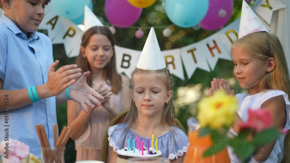 Girl blowing out candles on birthday cake as friends clapping rejoicing on holiday. Portrait of satisfied happy Caucasian child celebrating on backyard outdoors Girl blowing out candles on birthday cake as friends clapping rejoicing on holiday. Portrait of satisfied happy Caucasian child celebrating on backyard outdoors