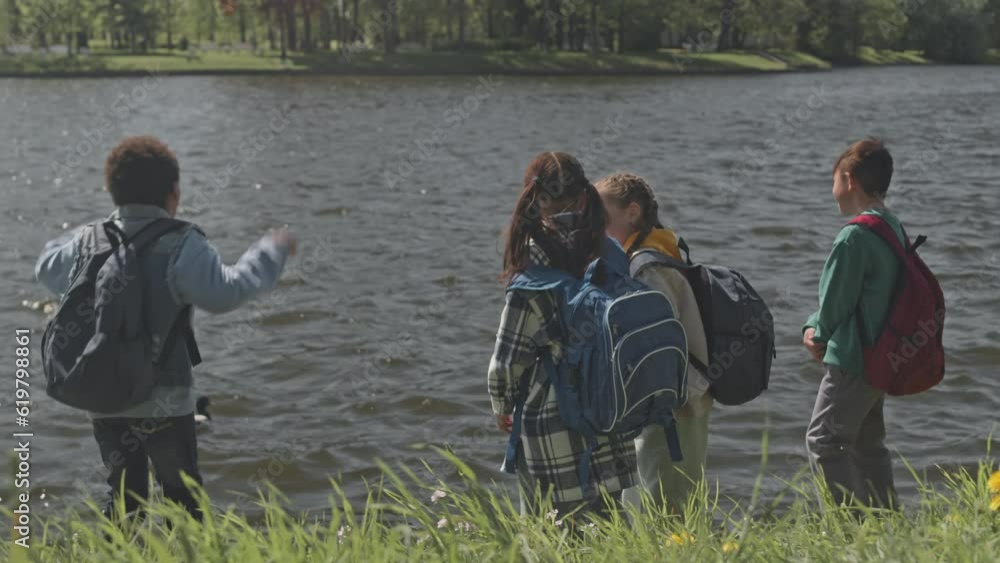 Back view of group of little kids standing in park and looking at duck swimming on pond while having walk together after school day Back view of group of little kids standing in park and looking at duck swimming on pond while having walk together after school day