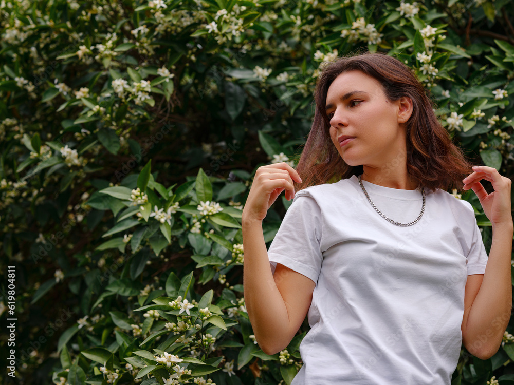 Beautiful young woman with dark curly hair and perfect skin wearing white tshirt posing near blossoming orange garden. Nude make up. Allergy season, antihistamines. Aroma oils natural perfumes Beautiful young woman with dark curly hair and perfect skin wearing white tshirt posing near blossoming orange garden. Nude make up. Allergy season, antihistamines. Aroma oils natural perfumes