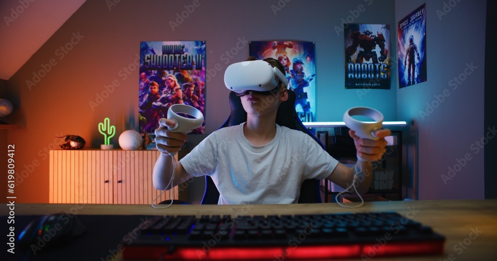 Young gamer in VR headset plays virtual online video game using wireless controllers. Boy enjoys eSports tournament. Room with warm neon LED lights. Gaming at home. POV from PC screen perspective. - obrazy, fototapety, plakaty 