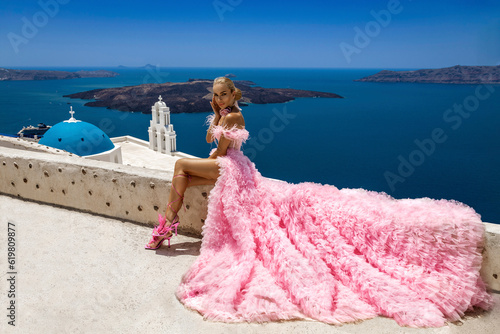 Glamour, stylish elegant bride woman in pink long wedding dress is posing near Church of the Three Bells in Fira in Santorini.