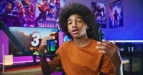 African American teenage gamer, blogger speaks on camera using microphone, shows thumbs up. Recording video games review for social networks. Neon cozy room. PC with online 3D shooter on background.