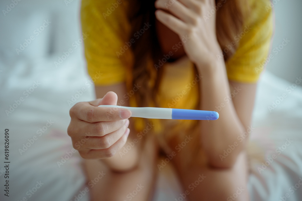 Obraz premium Young asian young woman feeling shock after look at positive Pregnancy test. Disappointed unplanned pregnancy girl sit on bed with upset and sad face for problem don't want to pregnant.