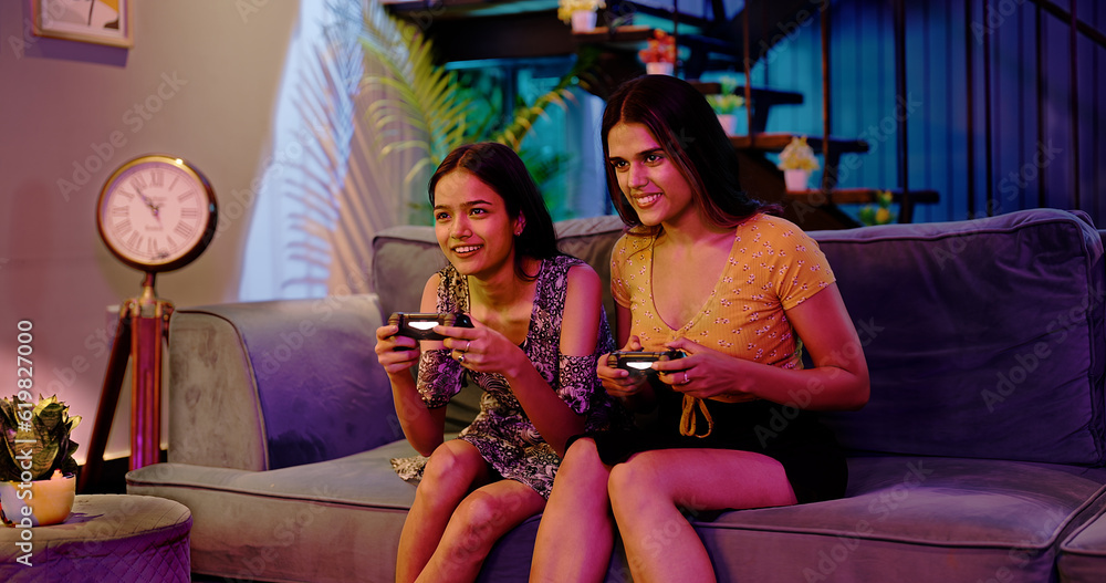 Obraz premium Happy Asian Indian two teen girl adult women couple playing video game hold joypad in hand sitting sofa couch making funny playful moment enjoy fun joy weekend late night at indoor interior home house