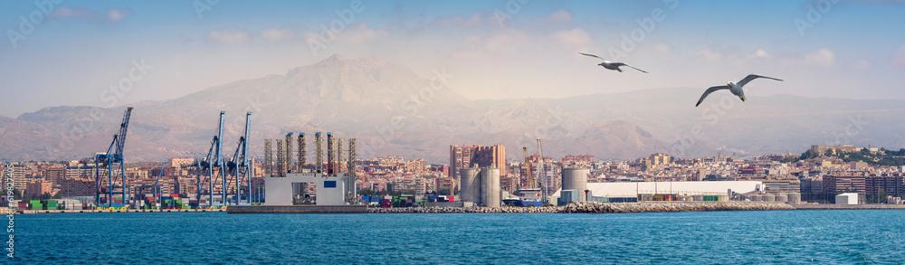 Obraz premium Industrial landscape of The Port of Alicante. Infrastructure of the seaport and cargo terminal with portal cranes and cargo containers storage. Global transportation hub on Mediterranean Sea in Spain.