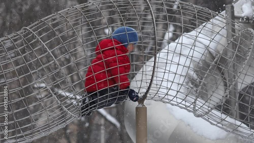 Winter walk with a small child on the playground.A kid in a winter jacket and hat climbs into the game tunnel.Outdoor activities for children in winter.In winter on the playground