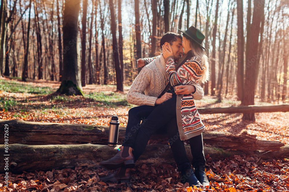 Portrait of young loving couple drinking tea from thermos relaxing in fall forest. Man and woman sit on trunk hugging Portrait of young loving couple drinking tea from thermos relaxing in fall forest. Man and woman sit on trunk hugging