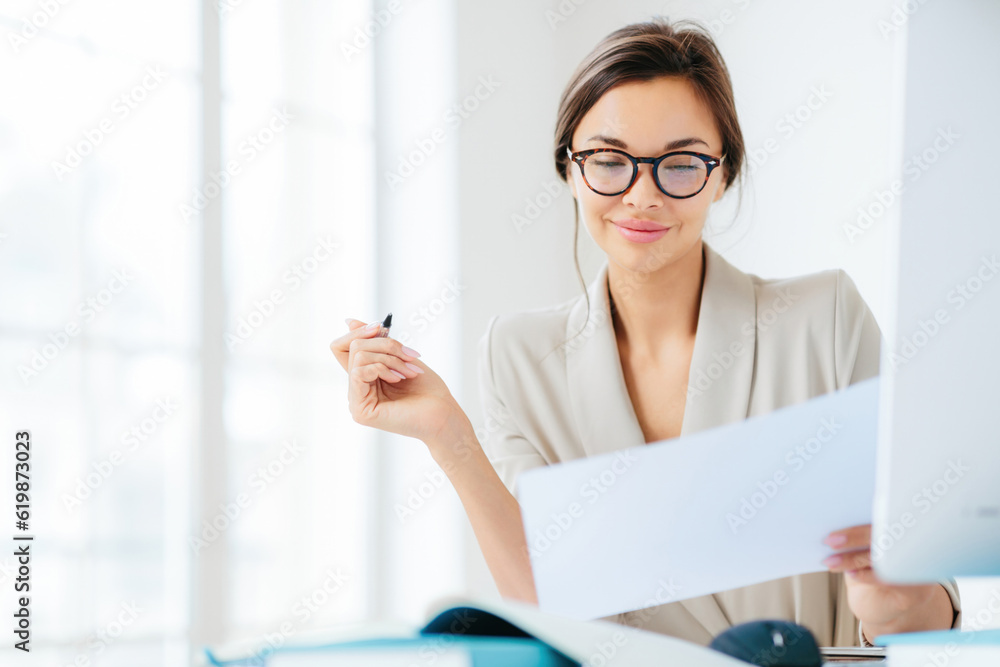 Concentrated businesswoman studies contract terms, writes with pen, dressed formally, at desk in spacious white interior Concentrated businesswoman studies contract terms, writes with pen, dressed formally, at desk in spacious white interior