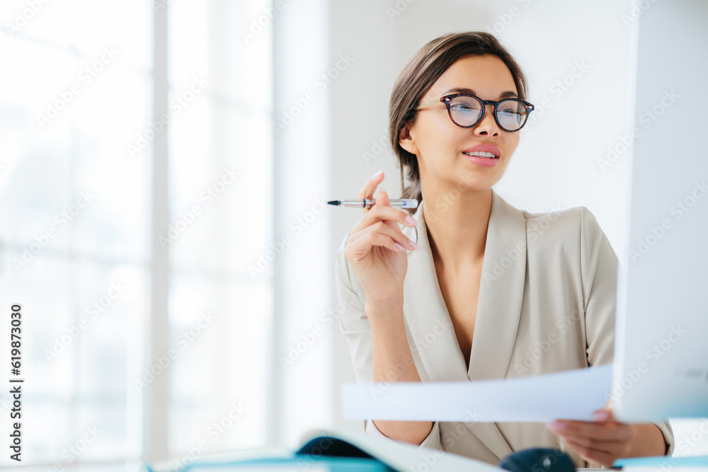 Focused businesswoman studies finances on computer, holds document and pen, works in office, wears formal attire and glasses Focused businesswoman studies finances on computer, holds document and pen, works in office, wears formal attire and glasses