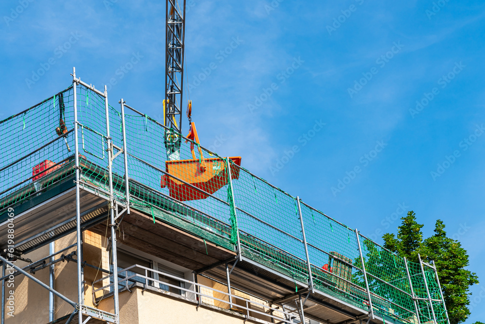 Lifting crane holds an orange container over the roof of a renovated house. Scaffolding green mesh enclose the roofl.