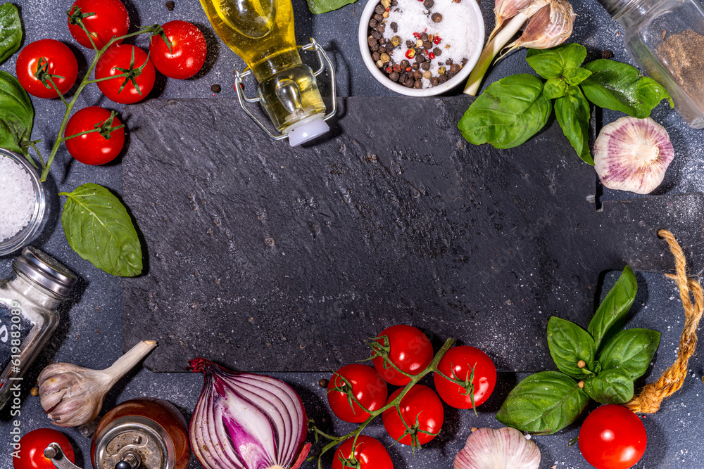 Black cooking background with useful cooking italian Mediterranean ingredients - tomatoes, basil leaves, greens, olive oil, salt, pepper, garlic, flat lay black concrete table top view copy space