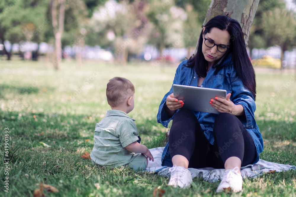 Business lady with smartphone and tablet sitting with little baby child in tree shadow in green park woman freelancer tries working online with son outdoors Business lady with smartphone and tablet sitting with little baby child in tree shadow in green park woman freelancer tries working online with son outdoors