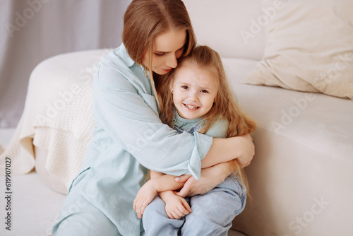 A young beautiful mother in blue home clothes and a little daughter are sitting on the floor in a cozy living room.