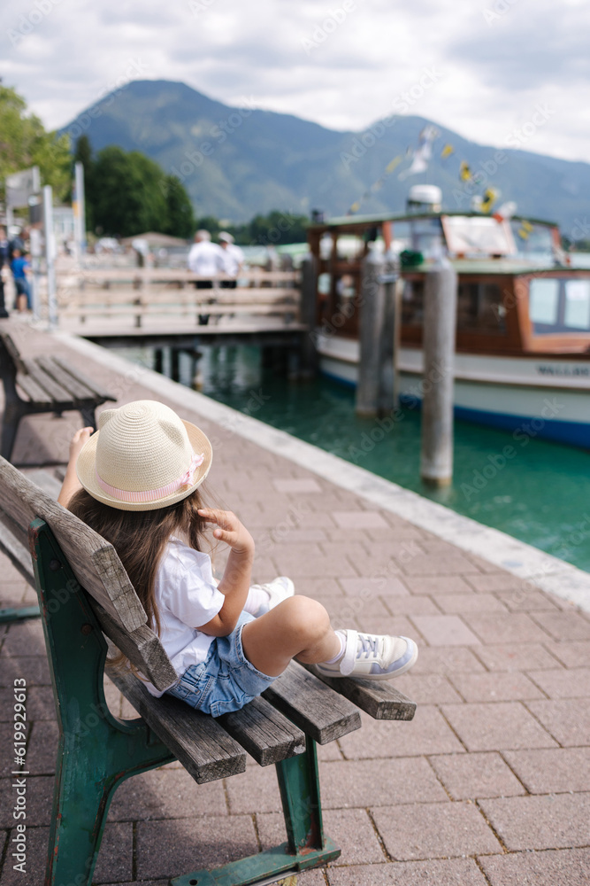 Back view of a little girl in a straw hat sitting on a bench and looking at a ship. Port