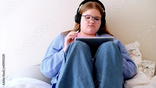 Relaxed teen girl in headphones completing online tasks on school website via modern tablet focused teenager in black-rimmed glasses studying online at home photo