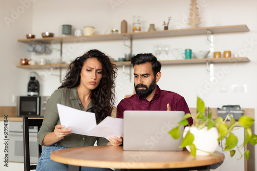 Bankruptcy concept. Overwhelmed arab couple sitting in kitchen in front of laptop, holding documents, got mail from bank